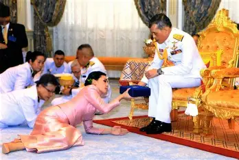 Royal Noble Consort Sineenat Wongvajirapakdi in pink attire kneeling and presenting a blue ceremonial item to King Maha Vajiralongkorn (Rama X), seated on a golden throne in decorated uniform, during her investiture at the Grand Palace, Bangkok, 28 July 2019, with attendants in white observing.