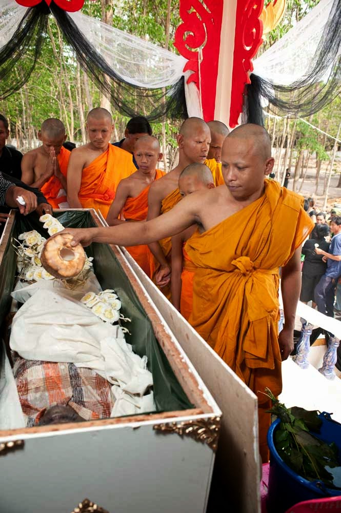 Colour image of an open casket containing the body of Seri, a Thai university graduate who died young. Buddhist monks in saffron robes sprinkle holy water over her body.