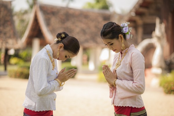 Two Thai women facing each other with hands pressed together and heads bowed in a wai greeting